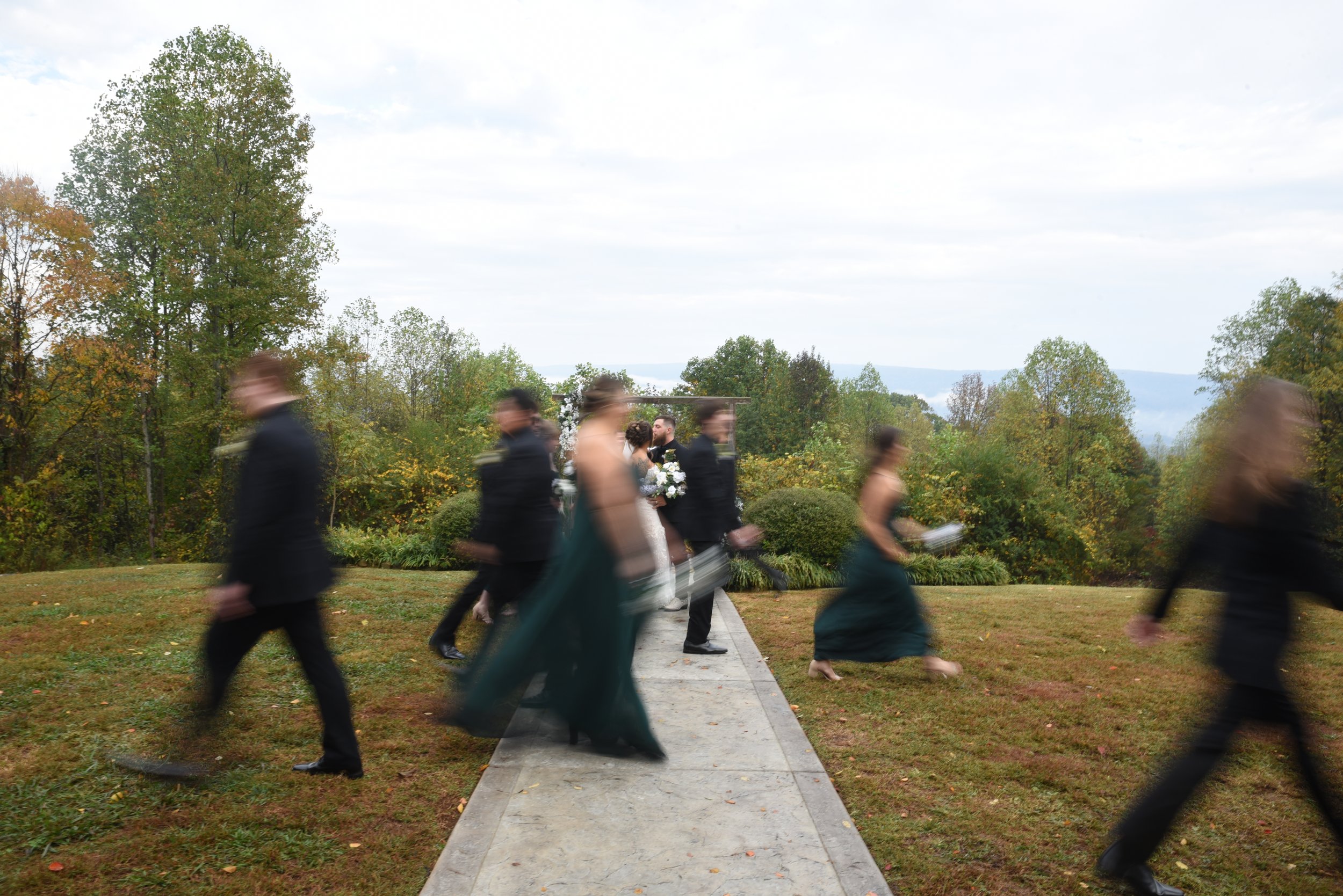 A bridal party walking past in motion blur as the bride and groom kiss at the altar arch, North Georgia hills in the background
