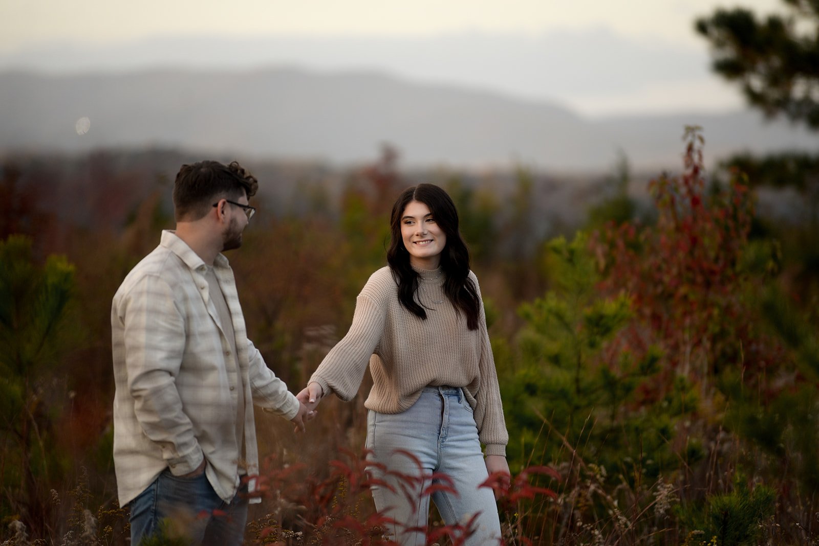 A North Georgia couple holding hands in tall autumn grass with mountain bokeh and fall foliage behind them — engagement photo outfit example
