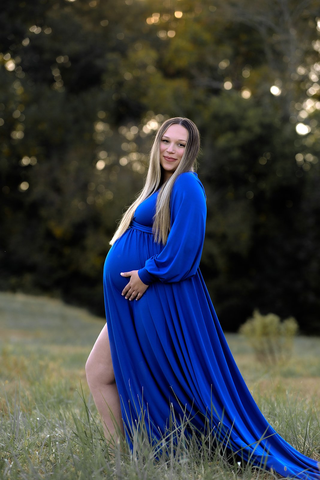 A pregnant woman in a long flowing electric-blue dress in a sun-dappled meadow at golden hour in Northwest Georgia — maternity styling reference